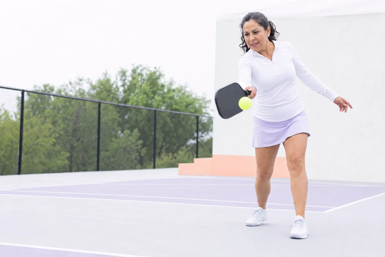 A woman in her late forties mid-swing on an outdoor pickleball court, paddle forward at the moment of contact, focused and enjoying herself