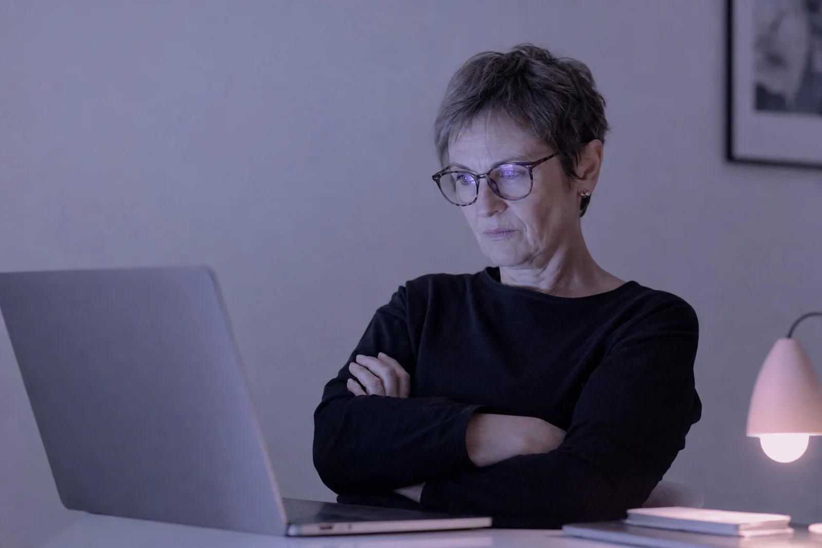 A woman in her fifties leaning back from her laptop at a home desk in the evening, arms folded, reading something on screen with a skeptical expression.