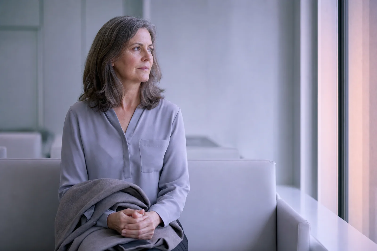 A woman in her fifties sitting in a quiet clinic waiting area, coat folded on her lap, looking toward a window with a composed expression.