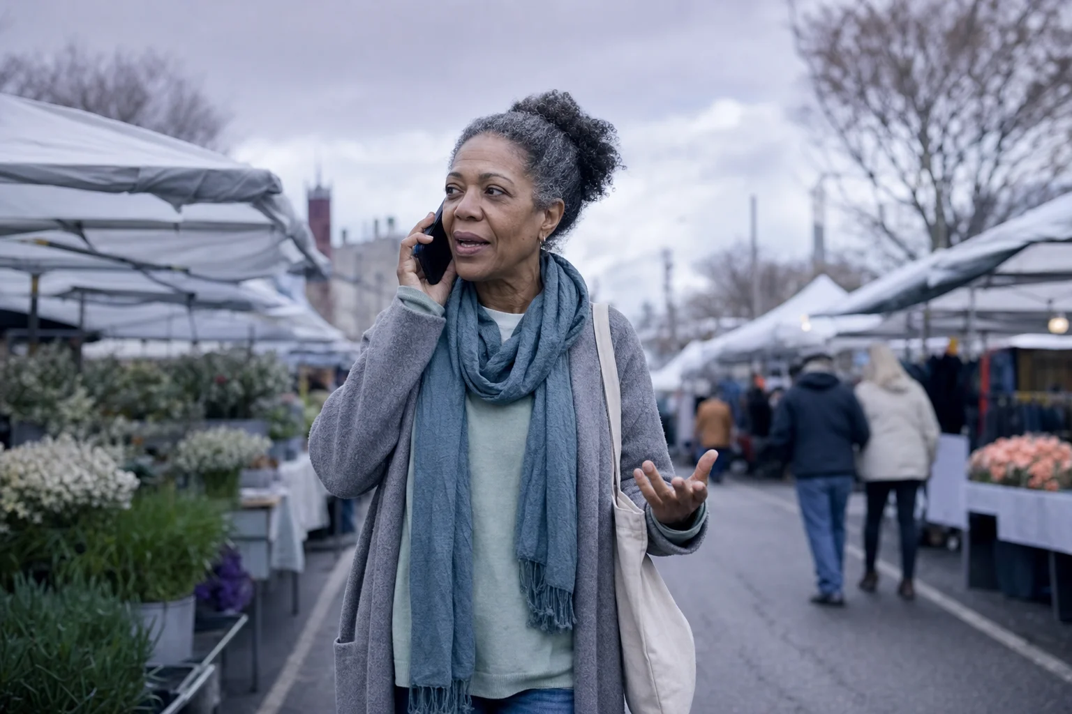 A woman in her fifties walking through an outdoor market on a phone call, tote bag on her shoulder, gesturing as she talks.