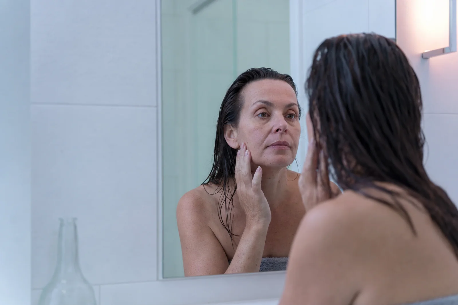 A woman in her fifties at a bathroom mirror with bare skin and damp hair, fingertips at her cheekbone, appraising her reflection frankly.