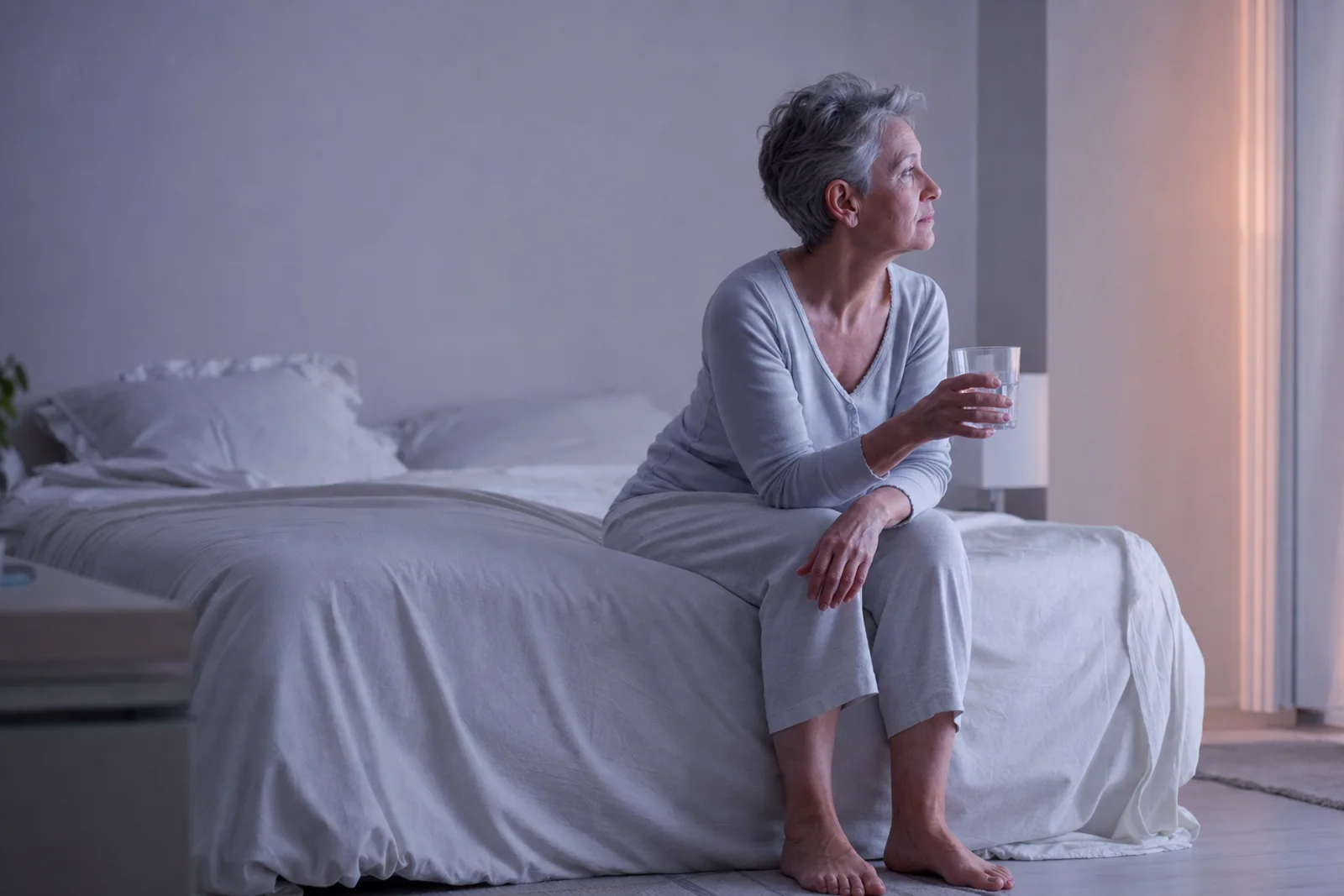 A woman in her mid-fifties sitting on the edge of her bed in early morning light, holding a glass of water, mid-thought.