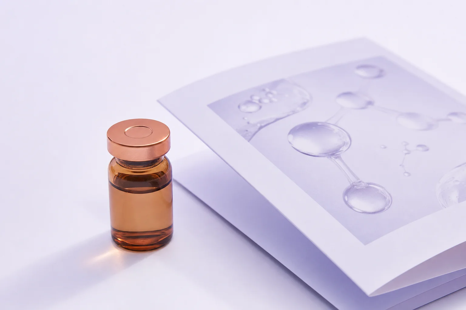 A small amber glass vial with a copper-colored cap beside a research paper on a near-white surface with cool lavender light