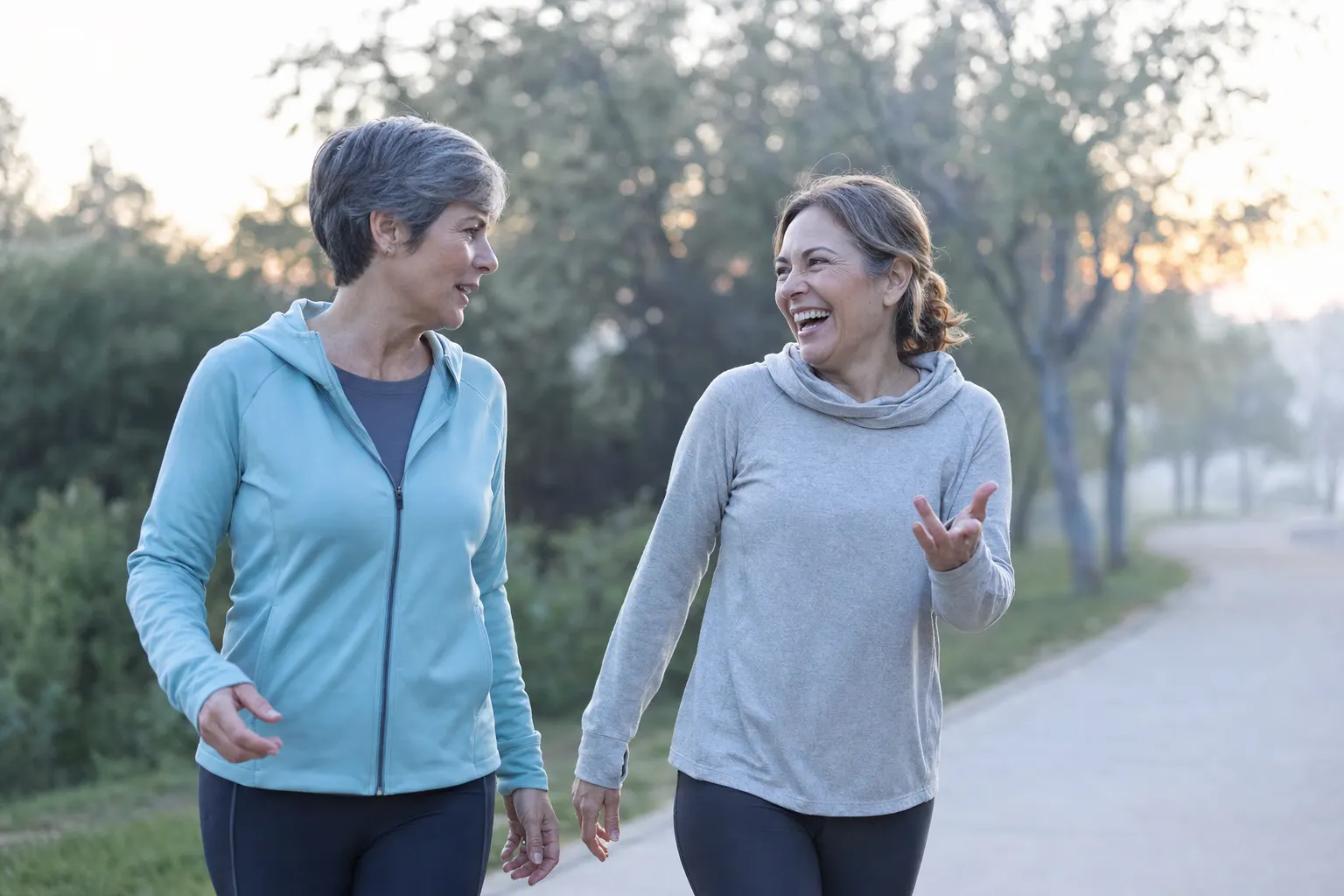 Two women in their fifties walking briskly along a park path, laughing mid-conversation, soft cool morning light