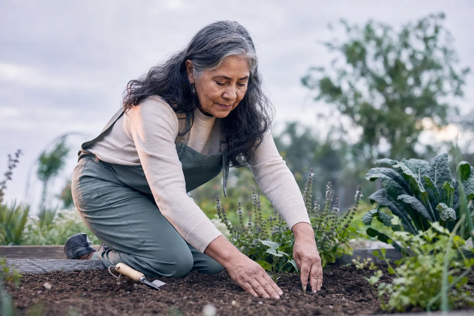 A Latina woman in her sixties kneeling in a garden bed, hands in soil, trowel beside her, soft cool daylight
