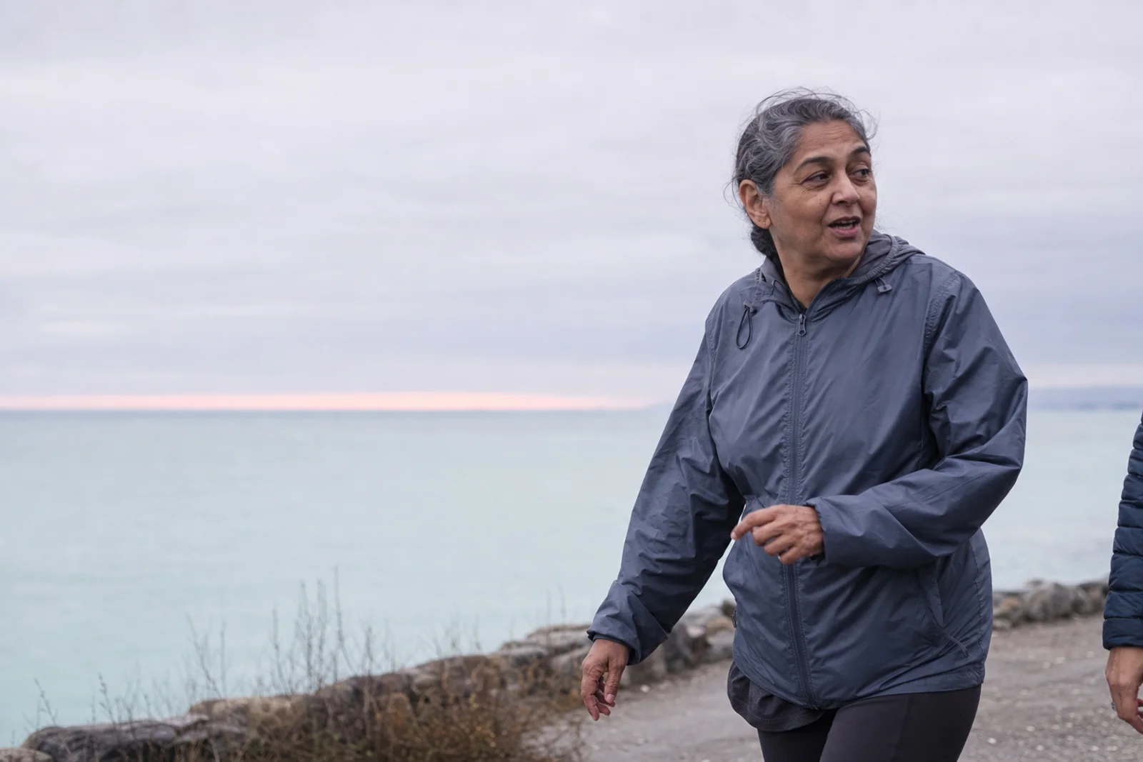 A woman in her fifties walking a coastal path on an overcast morning, mid-stride and mid-conversation