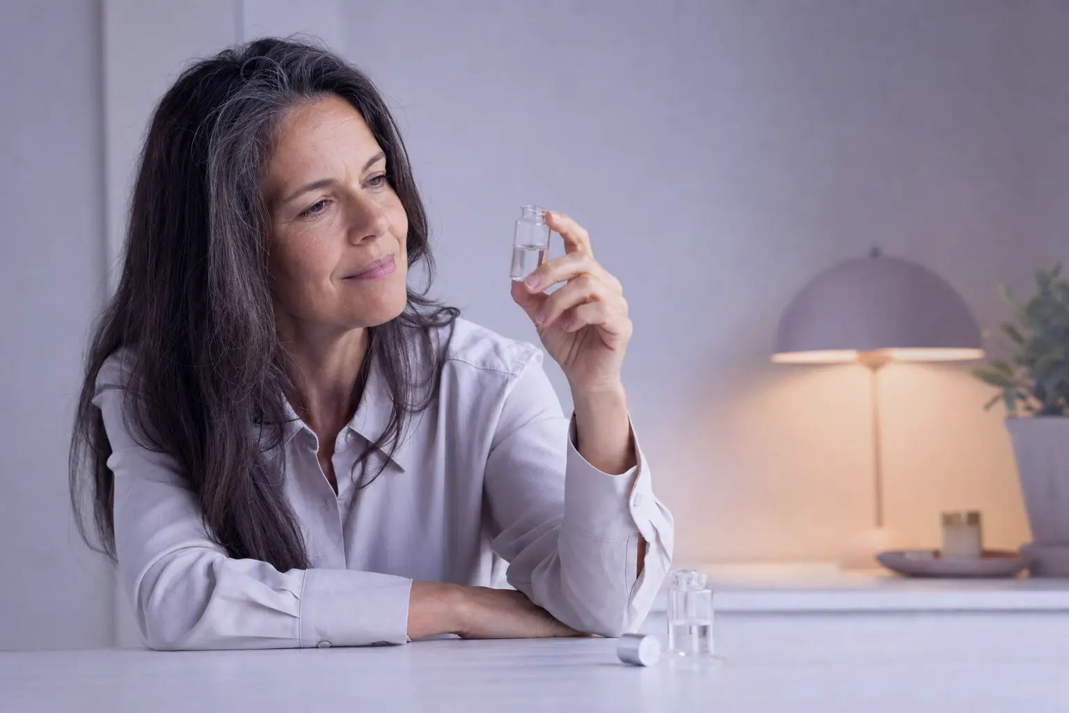A woman in her fifties studying a small glass vial at a near-white table in soft cool lavender light