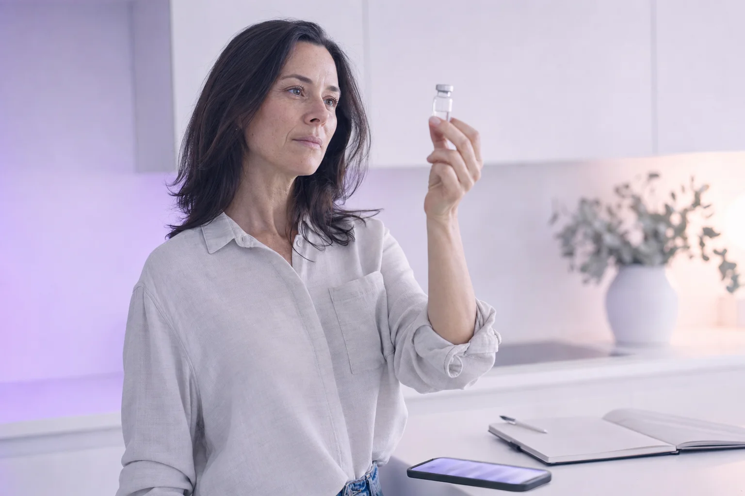 A woman in her forties at a near-white kitchen counter holding a small glass vial up to soft lavender light, considering it carefully before deciding