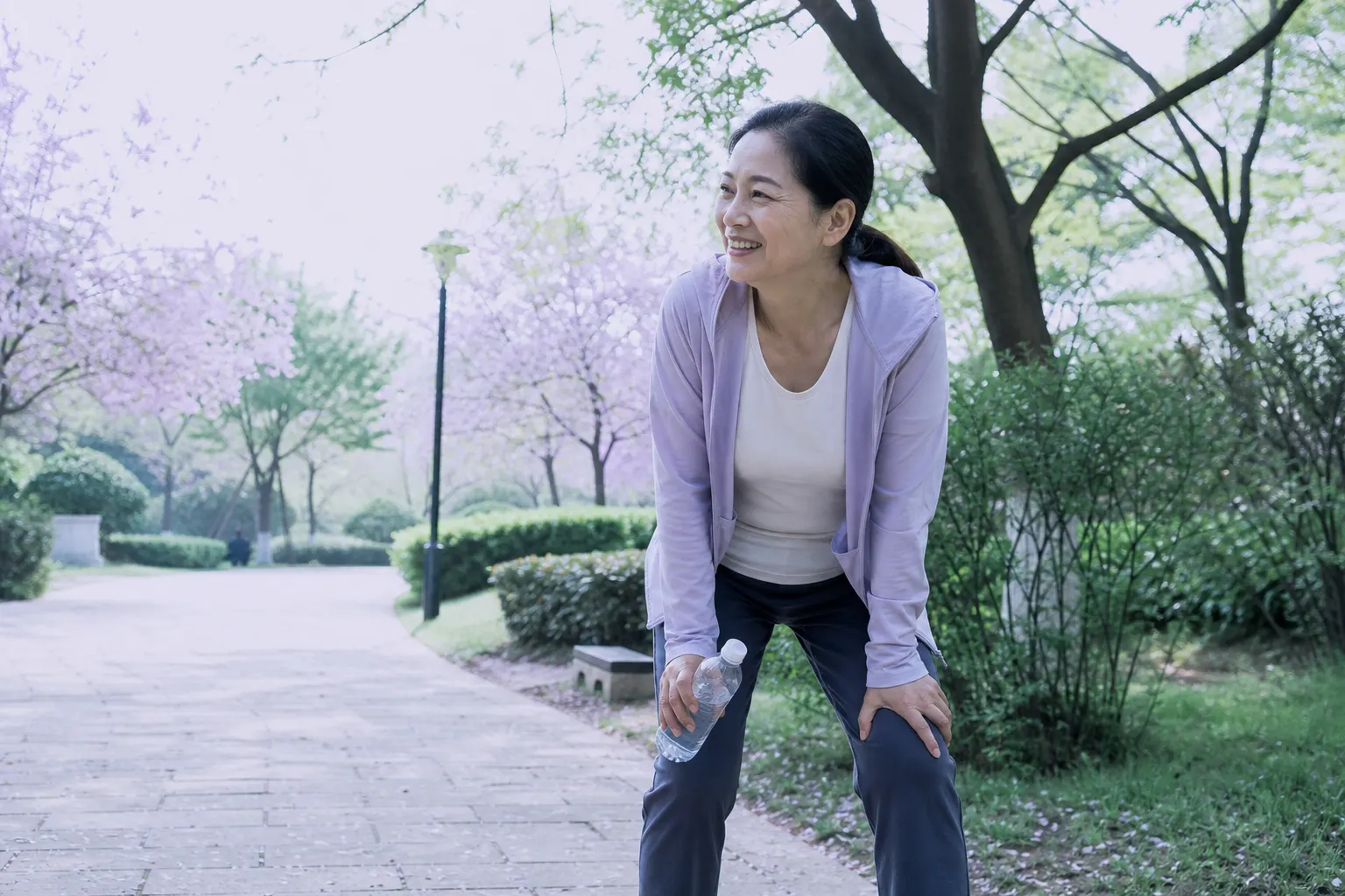 An East Asian woman in her mid-fifties in workout clothes catching her breath and smiling after a brisk walk in a bright park, water bottle in hand