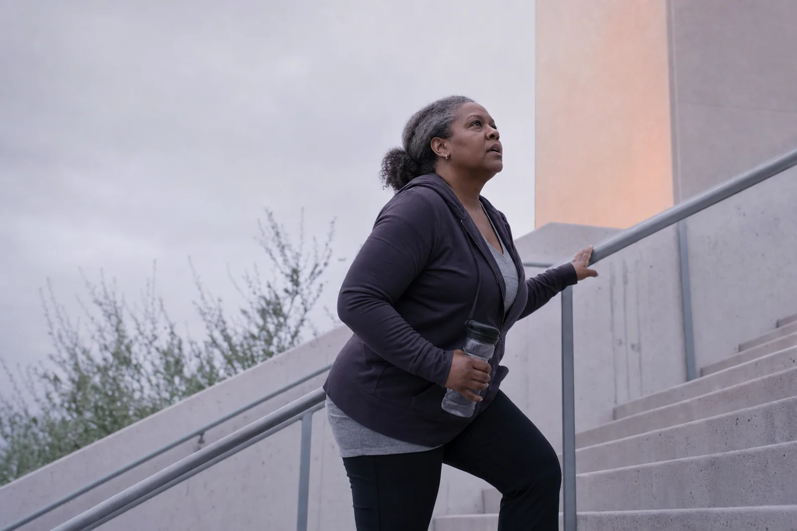 A woman in her fifties mid-stride on an outdoor staircase in cool morning light, one hand on the rail, moving with purpose