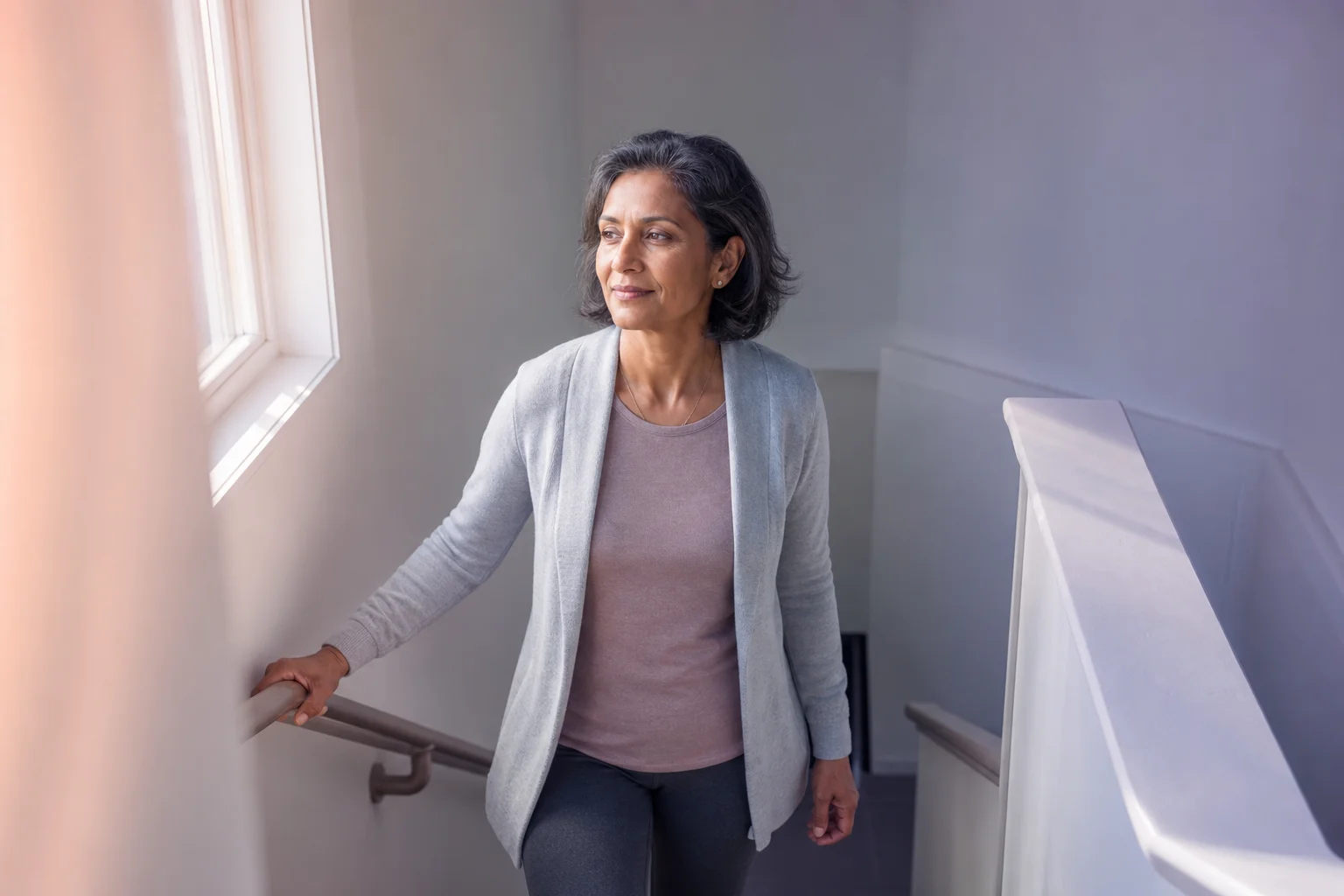 A South Asian woman in her mid-fifties pausing at the top of a sunlit stairwell, one hand resting on the railing, looking calmly out a window