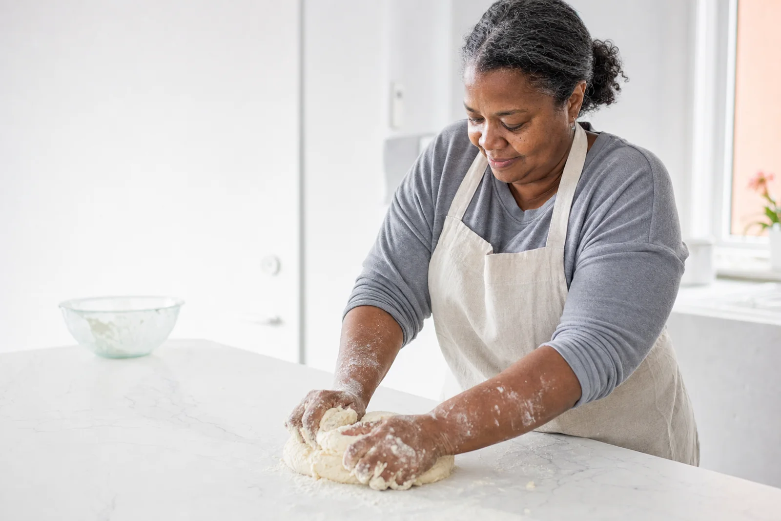 A woman in her fifties kneading dough at a kitchen counter in cool morning light, sleeves pushed up, mid-motion